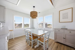 A dining area with a white table and chairs, a wicker pendant light, and a view through large windows.