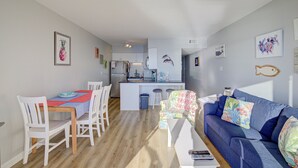 A modern living room with a blue sofa, white chairs, a wooden coffee table, and a kitchen area in the background. - Topsail Dunes condo - ocean-view balcony - beach access & beachfront pool (North Topsail Beach)