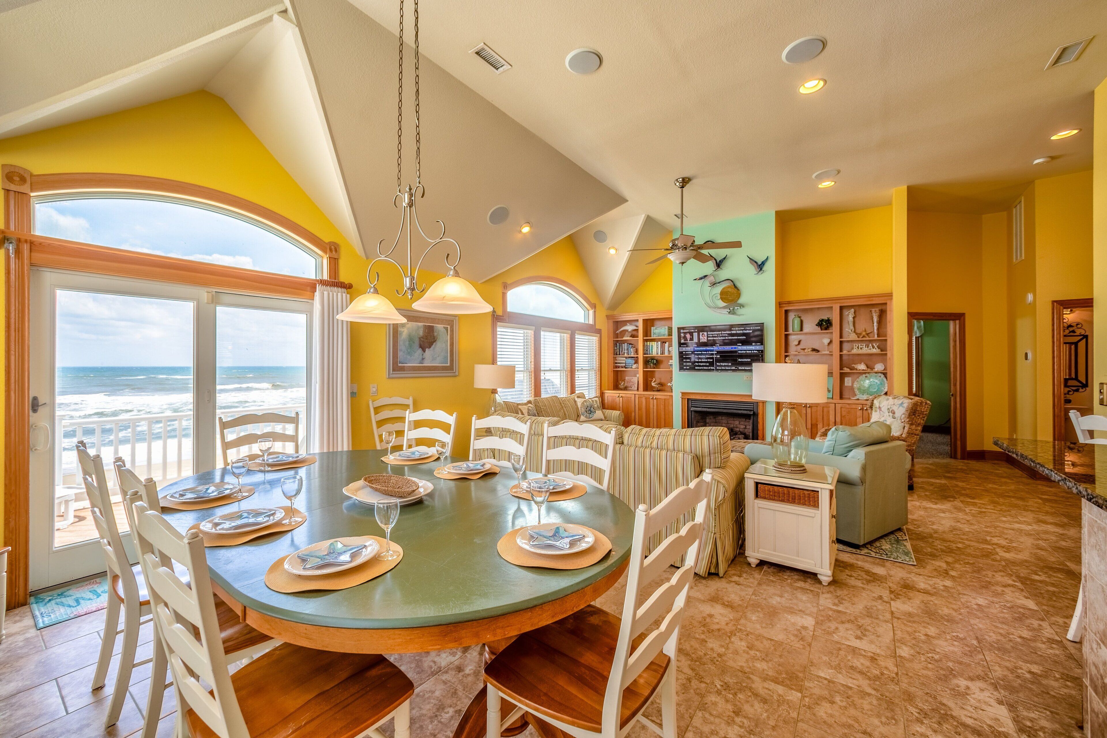 A dining area with a large table set for six, surrounded by white chairs, a kitchen area with a stove, and a view of the ocean through large windows.