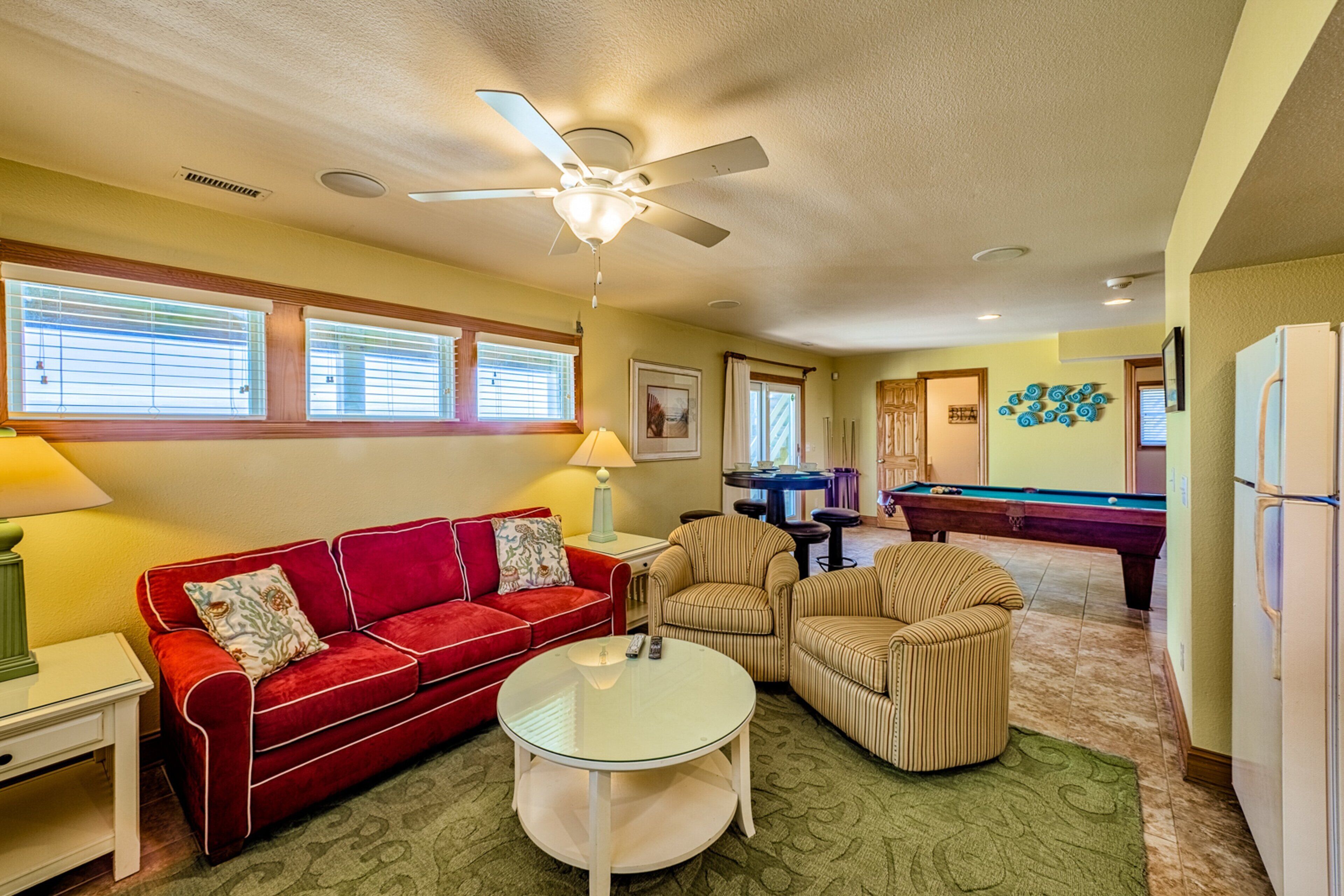 A living room with a red sofa, striped armchairs, a glass coffee table, a ceiling fan, and a pool table.