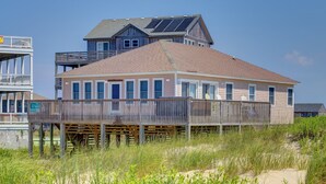 Una casa sulla spiaggia con veranda in legno, pannelli solari e un cartello con la scritta 'Affittasi'.