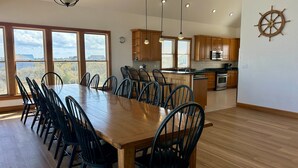 A spacious dining area with a long wooden table and black chairs, a kitchen with wooden cabinets and a ship wheel decoration, and large windows offering a view. - Ocean-view home with central AC, pool, & hot tub (Buxton)