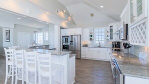 A spacious kitchen with white cabinetry, a central island with bar stools, stainless steel appliances, and a dishwasher. - Beachfront, multi-level home with pool, beach access & private hot tub (North Topsail Beach)