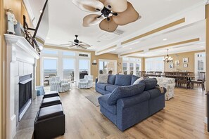 A spacious living room with a fireplace, a large blue sectional sofa, a ceiling fan, and a dining area with a chandelier. - Oceanfront home with volleyball, game room, pool, beach access & washer/dryer (North Topsail Beach)