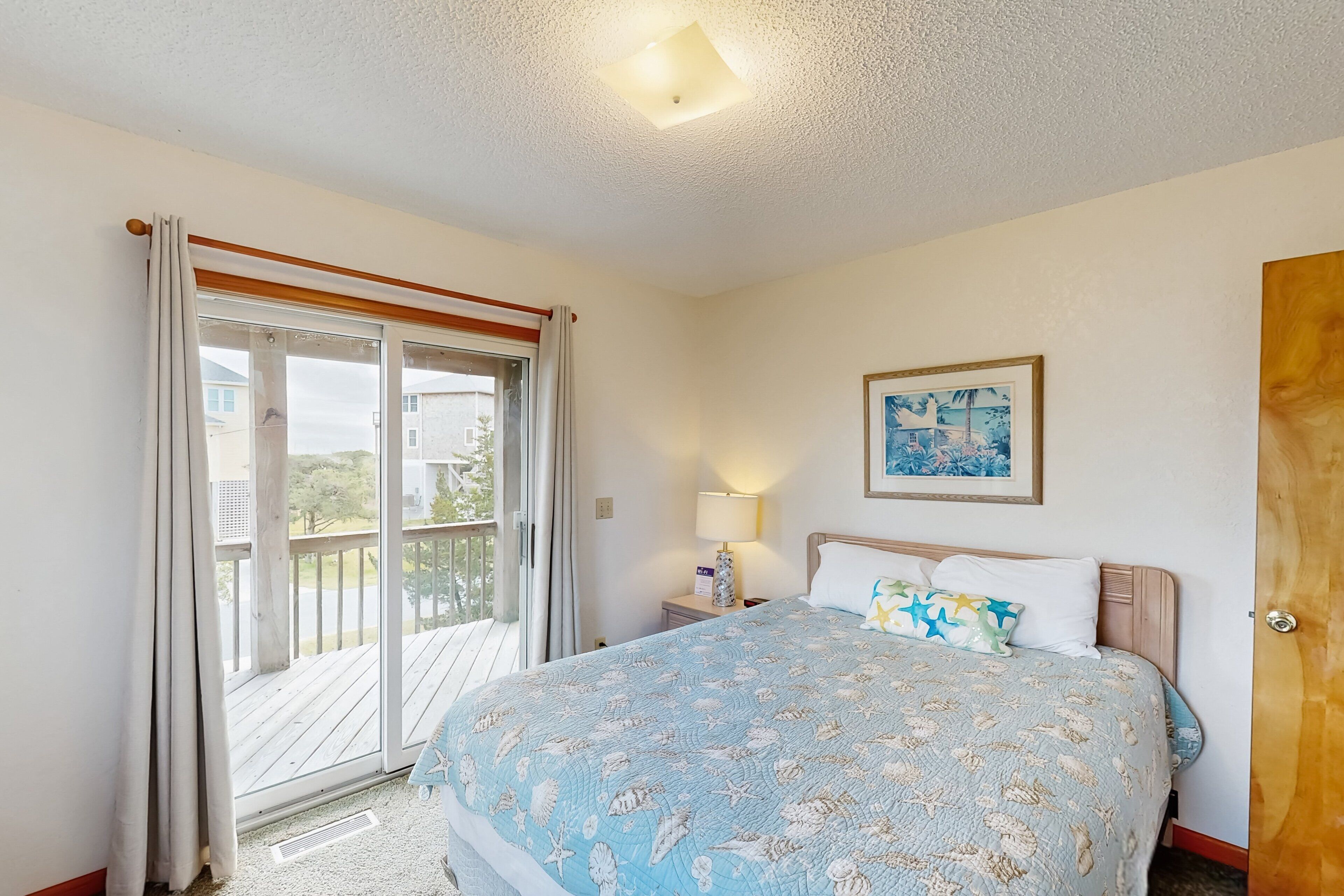 A bedroom with a bed, a nightstand, a lamp, a painting, and a sliding glass door to a balcony.
