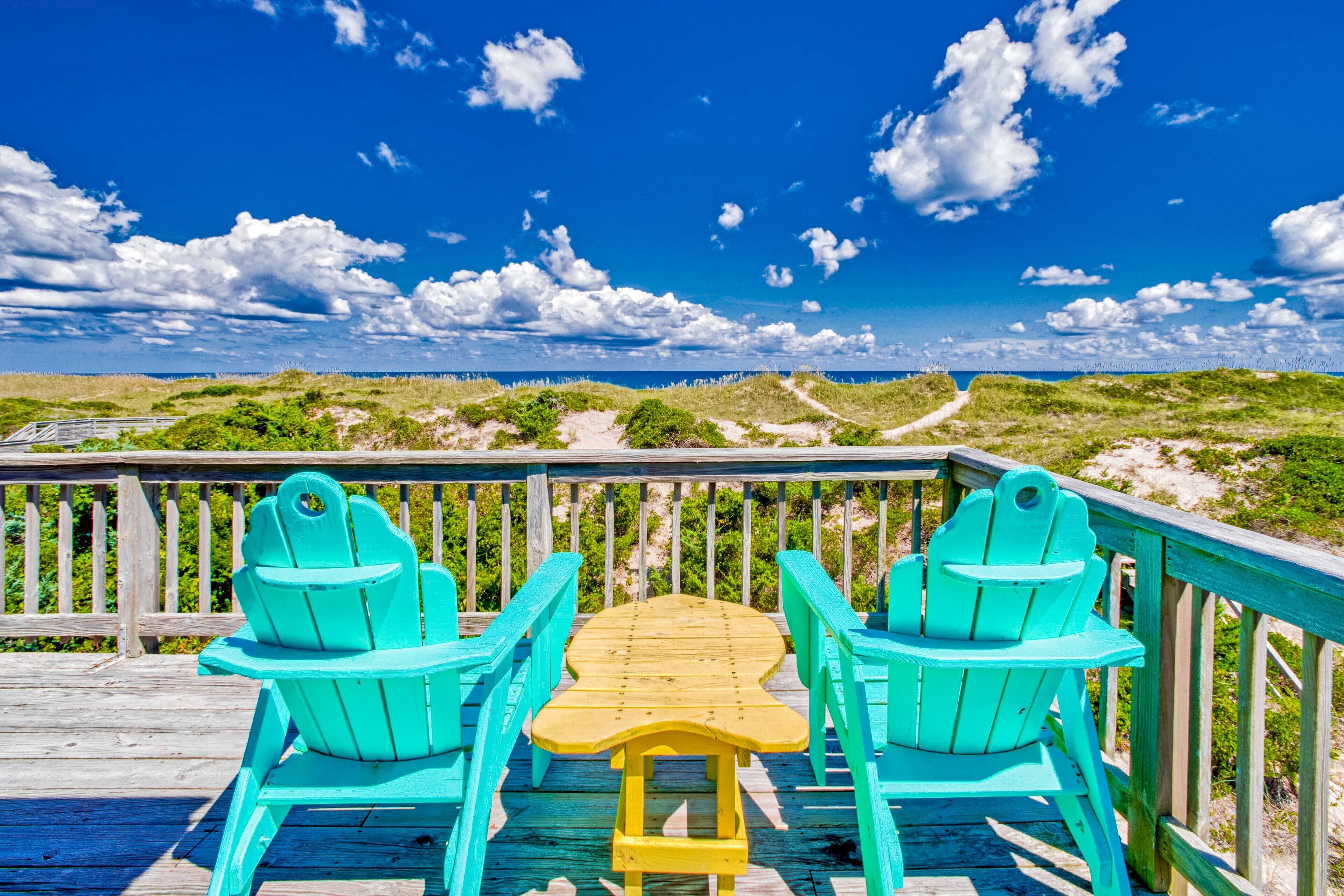 A wooden deck with two turquoise Adirondack chairs and a small table, overlooking a beach with dunes and a clear sky.
