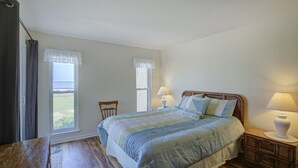 Ein Schlafzimmer mit einem großen Bett, zwei mit Vorhängen versehenen Fenstern, einem hölzernen Kopfteil und einem Nachttisch mit einer Lampe.
