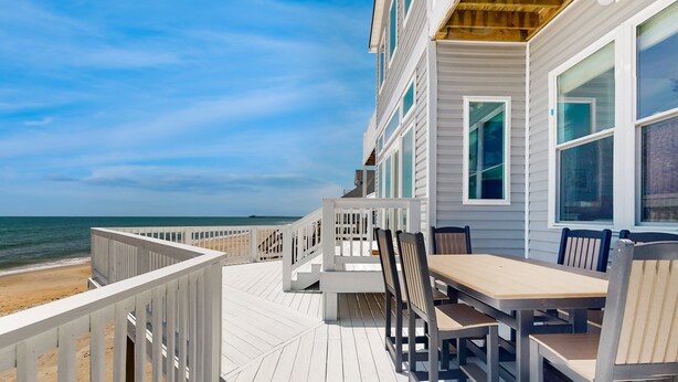 Een houten terras met een tafel en stoelen, uitkijkend op een strand.