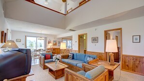 A spacious living room with a large flat-screen TV, wooden furniture, and a dining area with a chandelier. - Cape Joy 332: Lovely home 5 lots from beach w/screened porch (Avon)