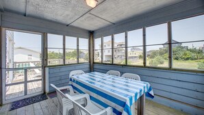 A covered outdoor dining area with a table and chairs, large windows, and a view of surrounding buildings. - Cape Joy 332: Lovely home 5 lots from beach w/screened porch (Avon)