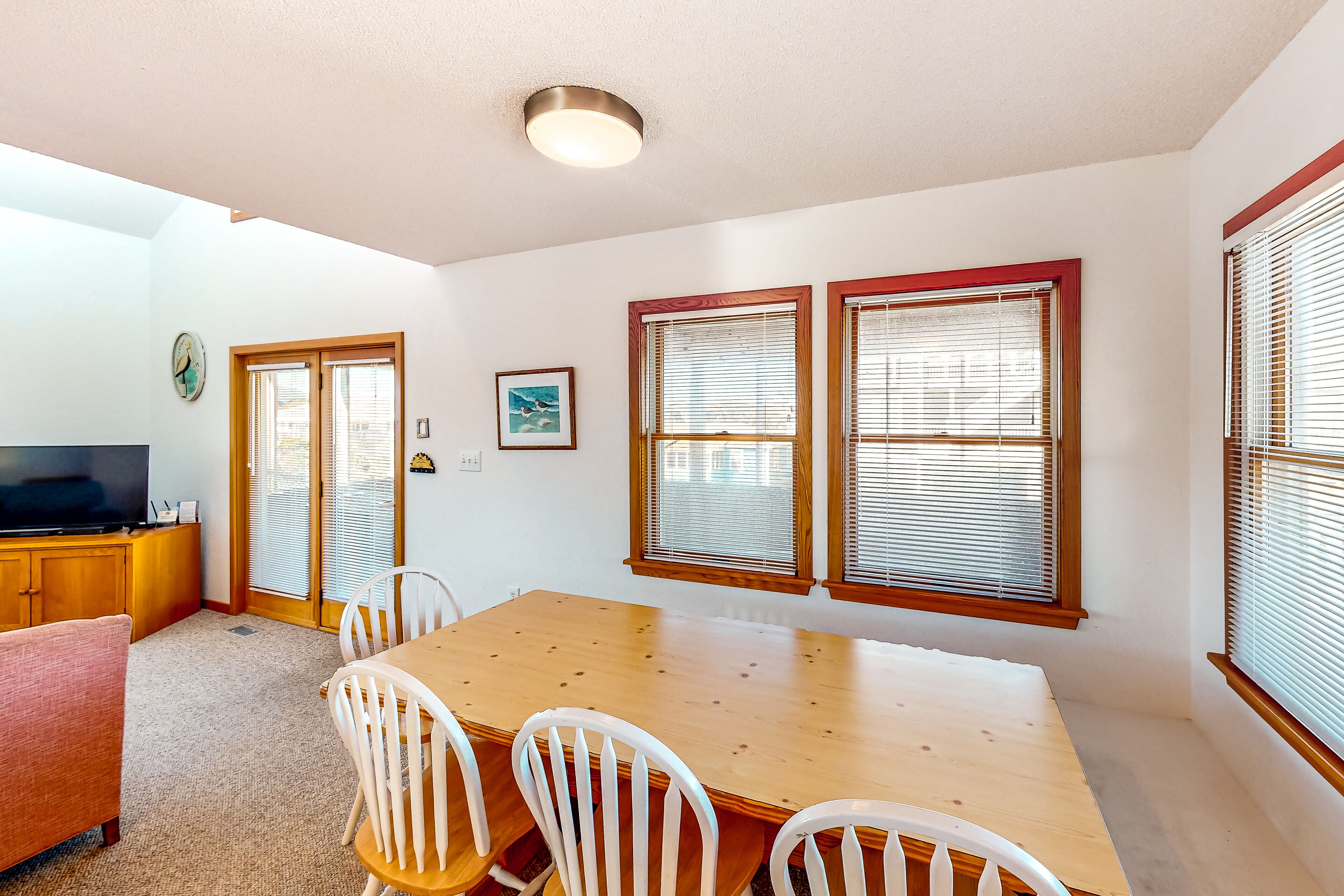A dining area with a wooden table and white chairs, a television on a wooden cabinet, and a window with blinds.