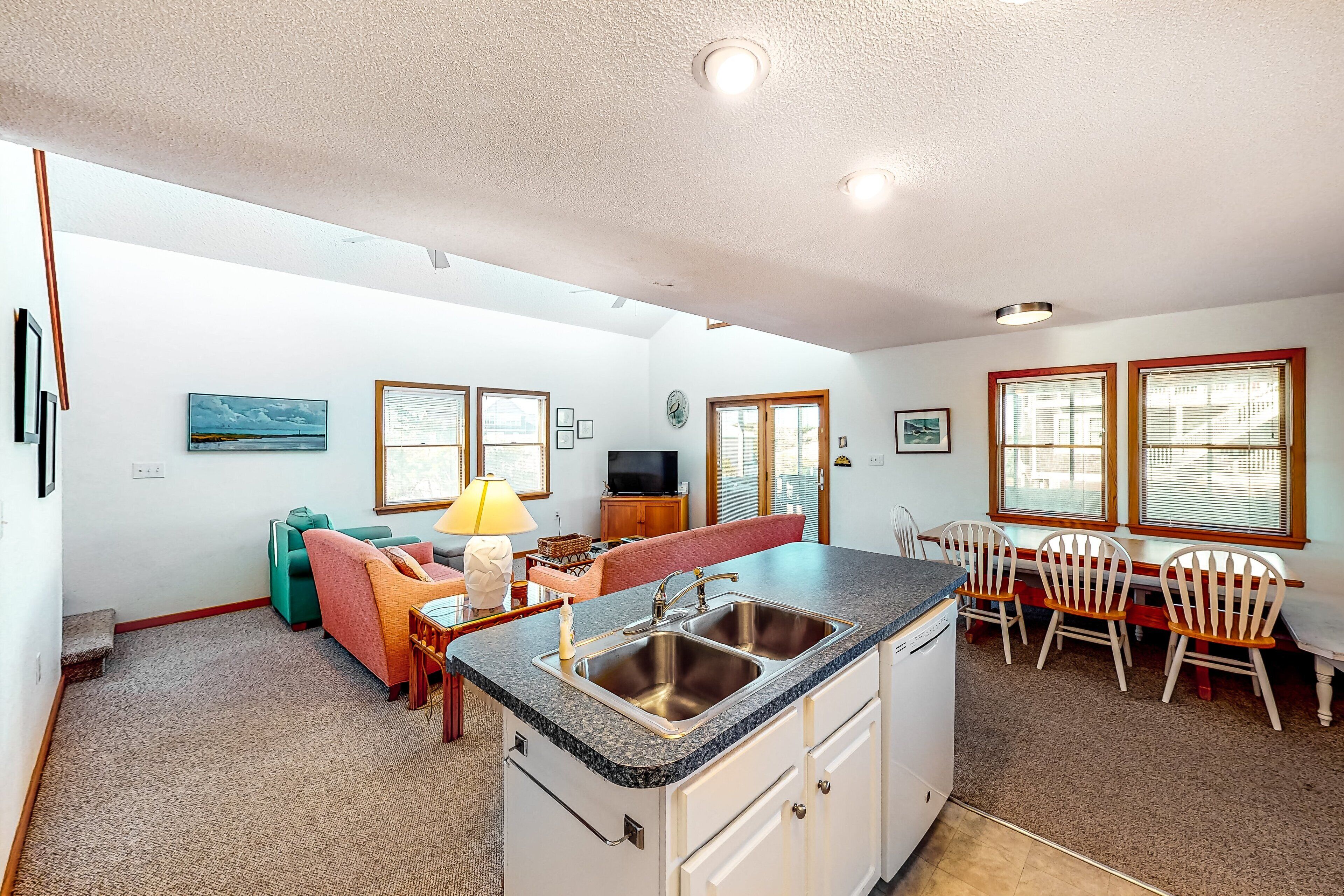 A spacious kitchen with a central island, a dining area with wooden chairs, and a living area with a sofa and a lamp.