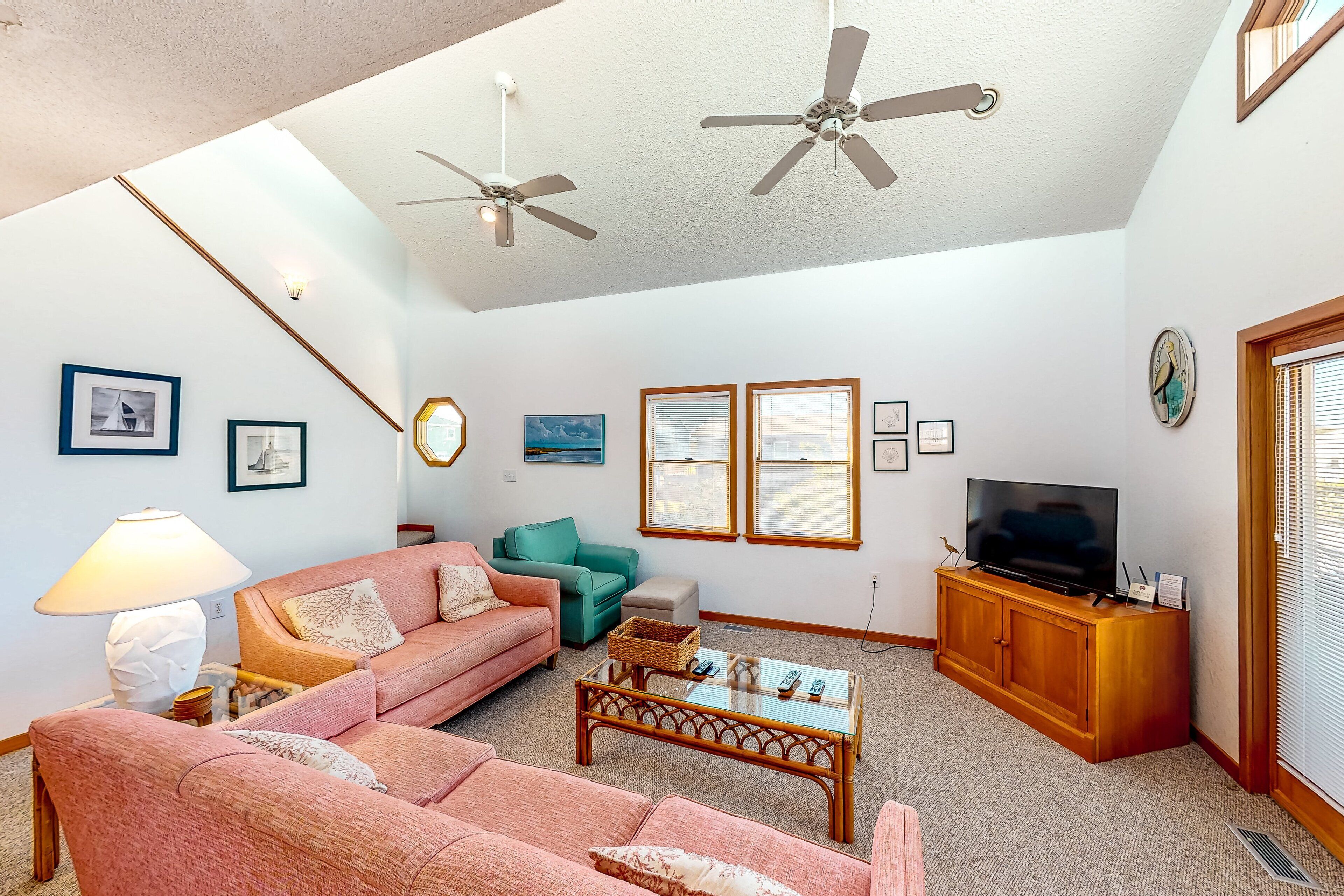 A living room with a pink sofa, teal armchair, and a coffee table. There are ceiling fans, a TV, and framed pictures on the walls.