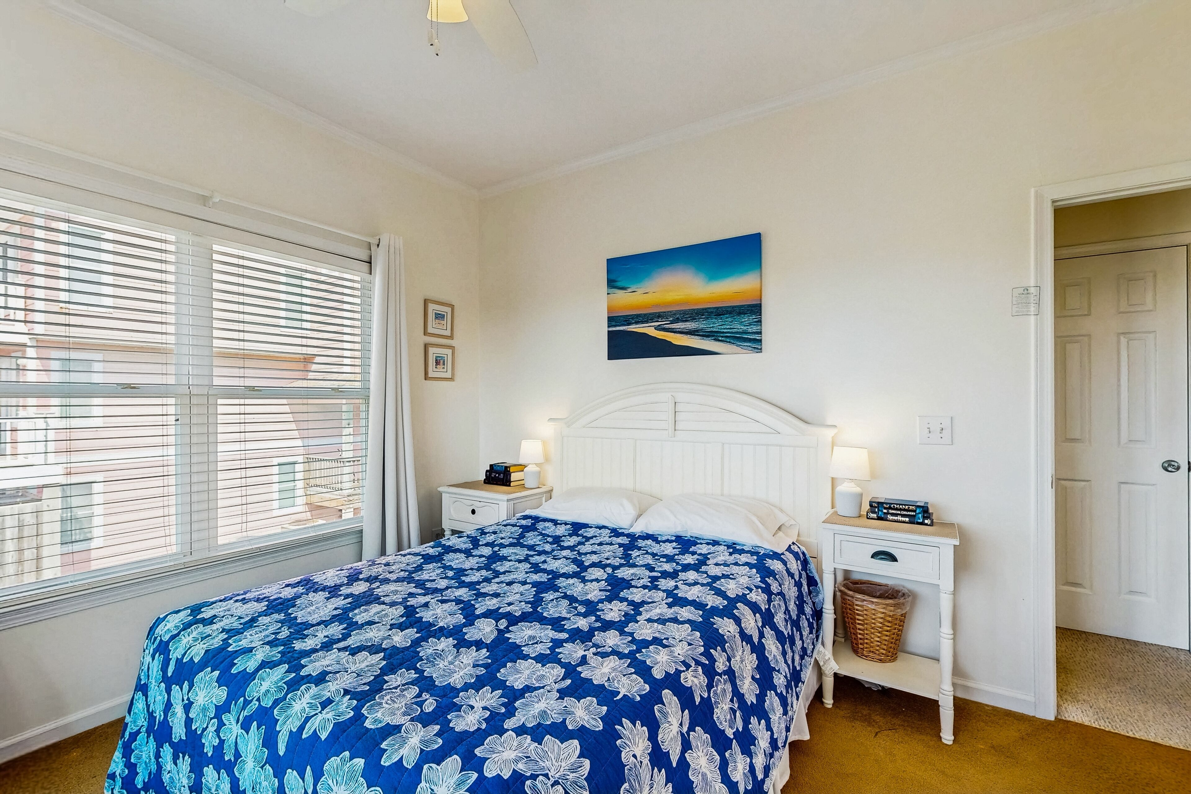 Un dormitorio con una cama grande, cabecera blanca, dos mesitas de noche con lámparas, un cuadro en la pared y una ventana con persianas.