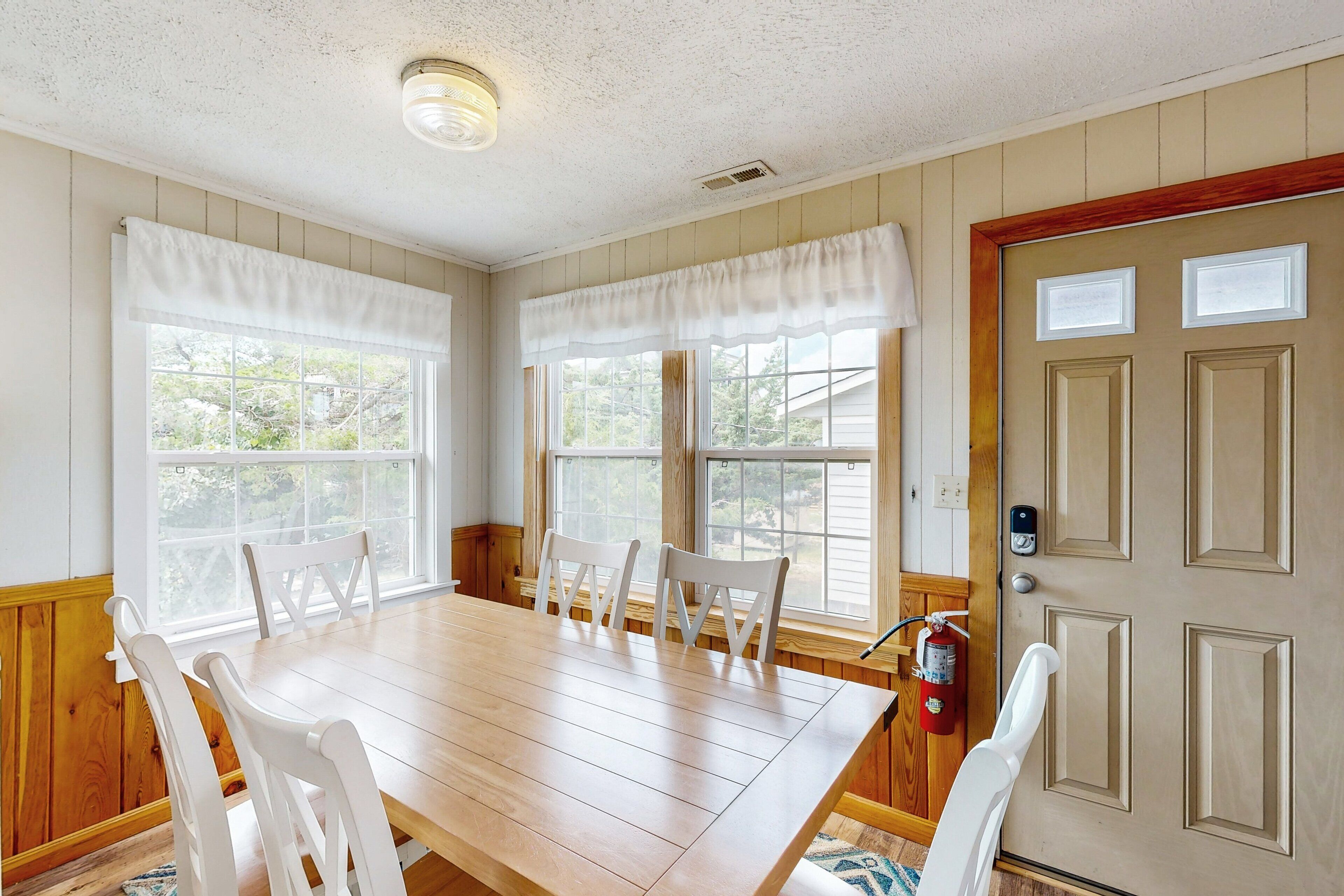 A dining room with a wooden table and white chairs, large windows with white valances, and a door with a fire alarm.