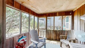 A wooden deck with two Adirondack chairs and a fan, large windows, and a view of trees and a house. - Pelican's Roost 467: Semi-oceanfront, dog-friendly, outdoor pool/tennis (Avon)