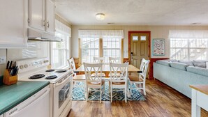 A kitchen with white cabinets, a stove, and a dining table with chairs. There is a window with a view of houses outside. - Pelican's Roost 467: Semi-oceanfront, dog-friendly, outdoor pool/tennis (Avon)