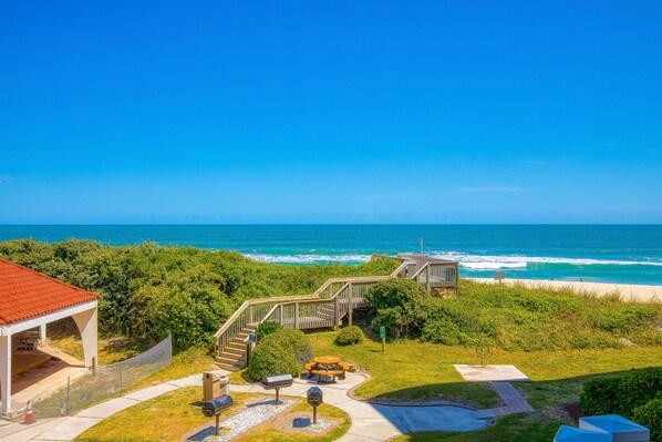 A coastal view with a wooden staircase leading to a beach, a covered area with outdoor seating, and a clear blue sky. - St. Regis beachfront condo with ocean & pool-view balcony, gym & sauna (North Topsail Beach)