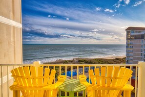 Un balcón con sillas Adirondack amarillas y una mesa verde, con vista a la playa y al océano.