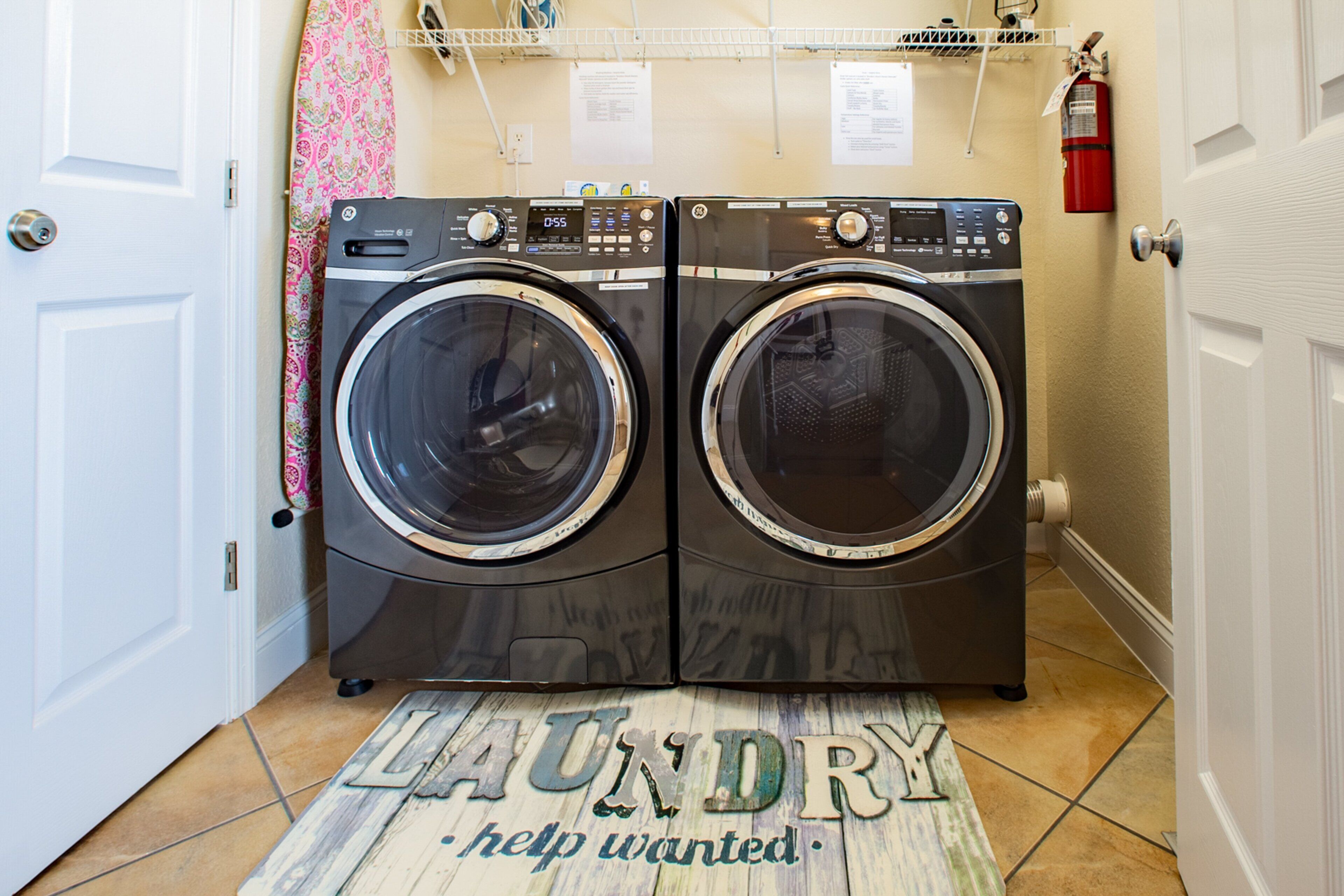 A laundry area with two front-loading washing machines, a fire extinguisher, and a laundry mat with the text "LAUNDRY help wanted."