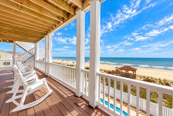 A wooden deck with white railings overlooking a beach with a gazebo and lounge chairs.