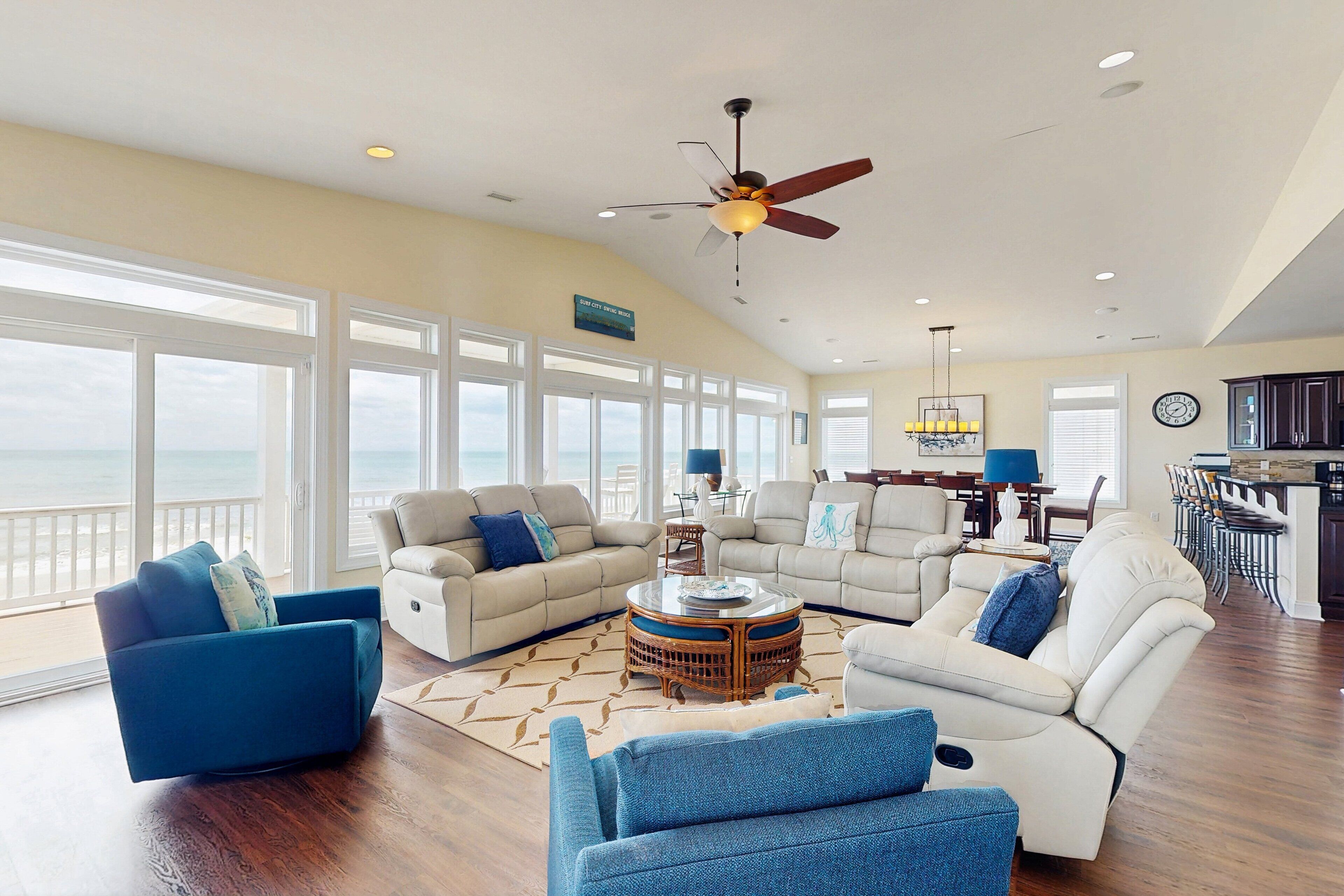 A spacious living room with a large window overlooking the ocean, a ceiling fan, and a dining area with a view.