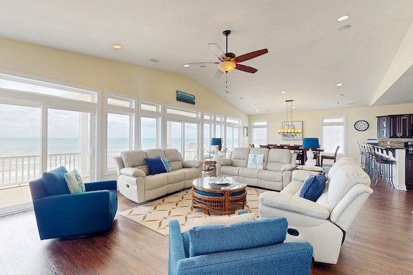A spacious living room with a large window overlooking the ocean, a ceiling fan, and a dining area with a view.