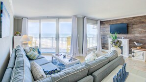Een woonkamer met een grote grijze bank, een salontafel en een televisie die aan een houten muur is bevestigd. Er is een balkon met uitzicht op de oceaan.