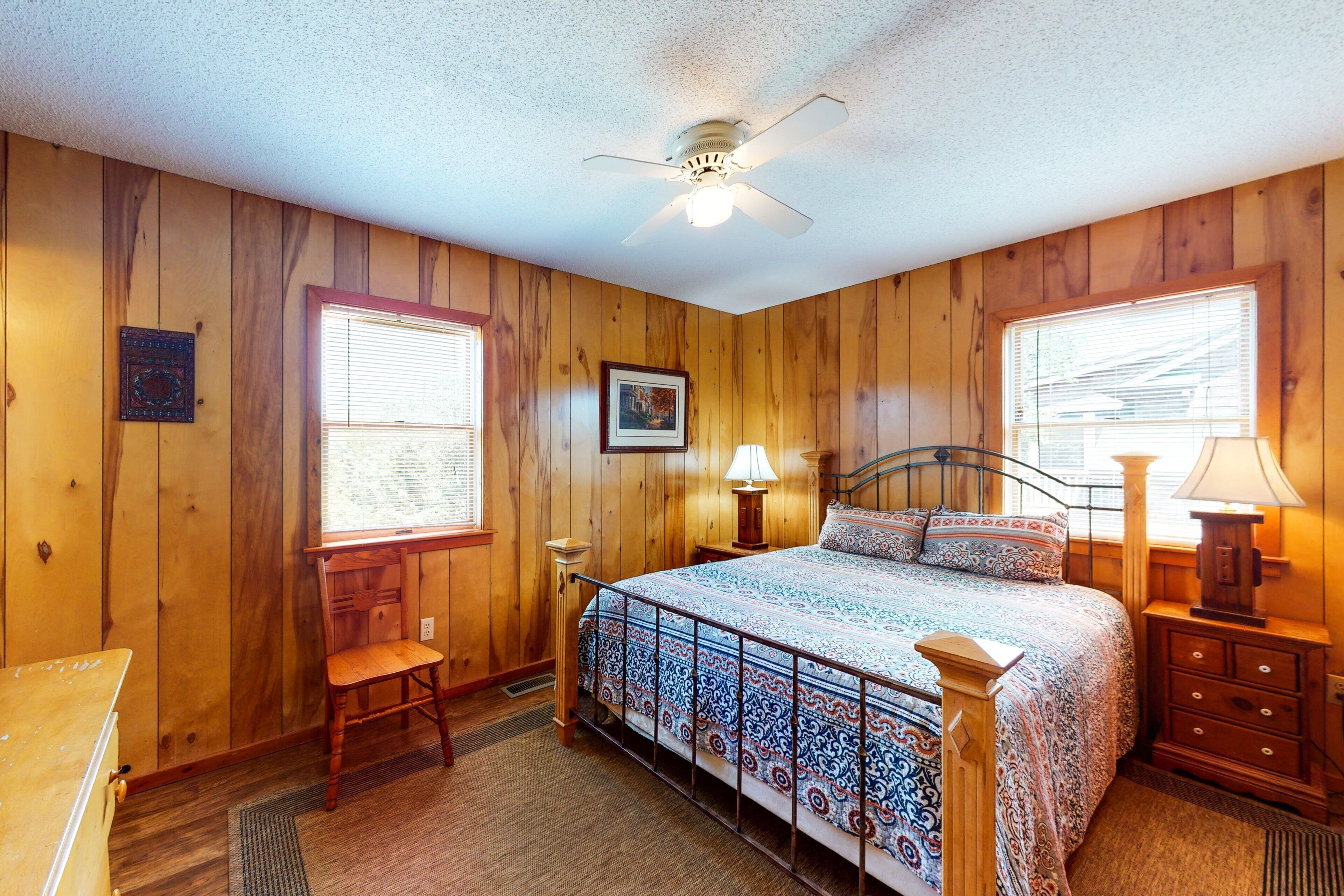 A bedroom with wooden walls, a bed with patterned bedding, a ceiling fan, and two windows with blinds.