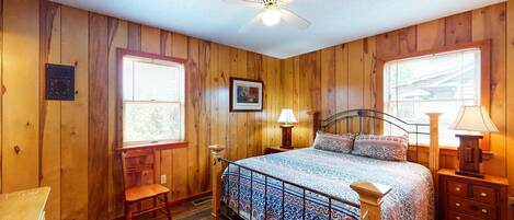A bedroom with wooden walls, a bed with patterned bedding, a ceiling fan, and two windows with blinds.