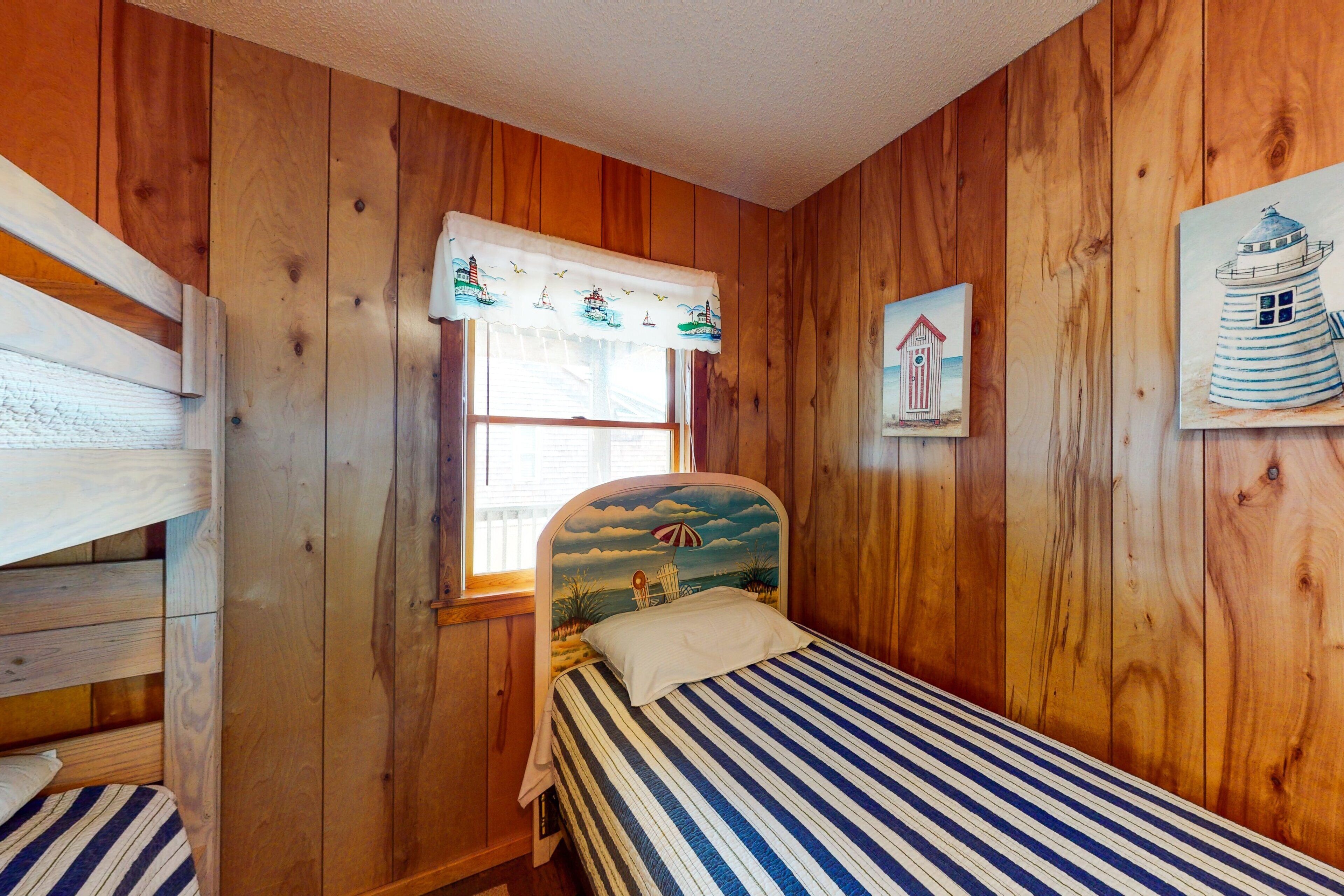 Een kamer met houten lambrisering, een bed met een hoofdeinde in strandstijl, een raam met uitzicht en een schilderij van een vuurtoren aan de muur.