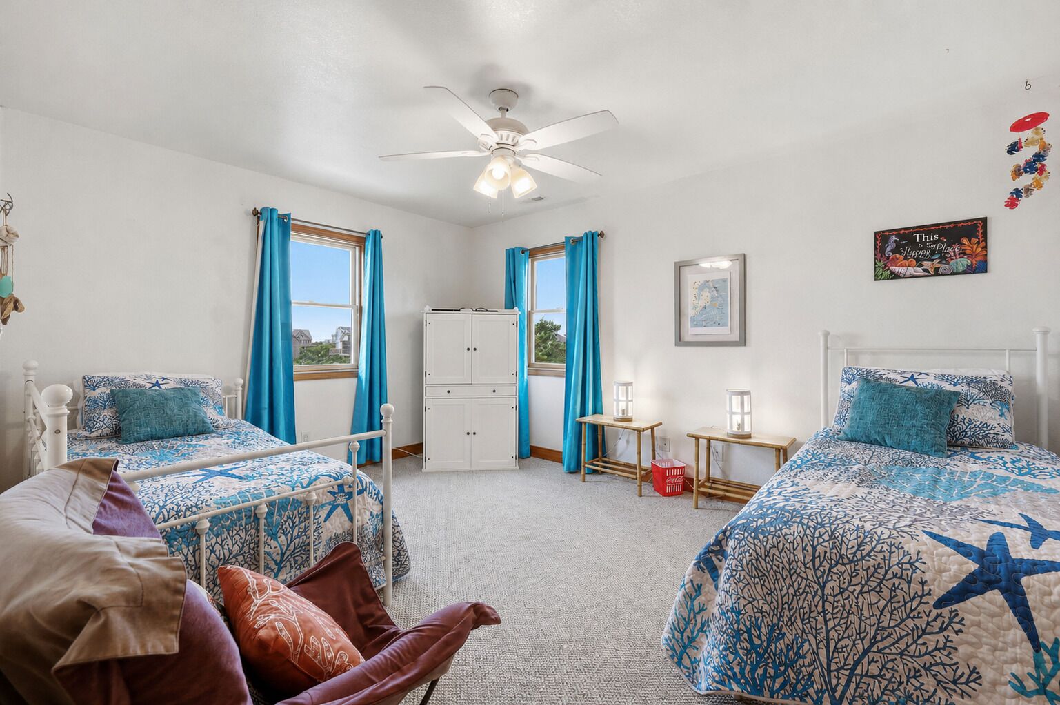 Une chambre avec deux lits, un ventilateur de plafond, une fenêtre avec des rideaux, une petite table avec une lampe et un tableau accroché au mur.