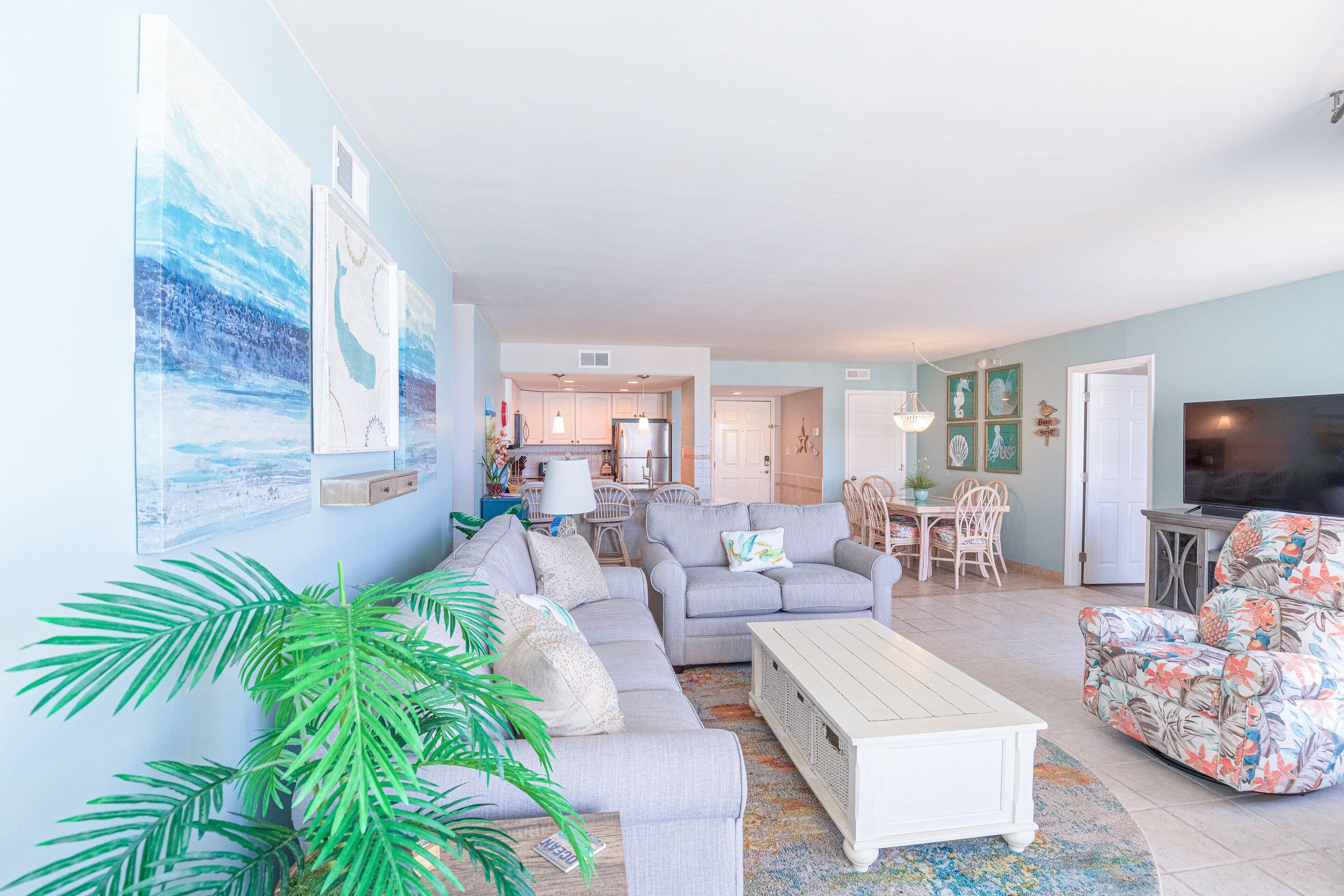 A spacious living room with a sectional sofa, a coffee table, and a TV. There is a dining area with a table and chairs, and a kitchen in the background.