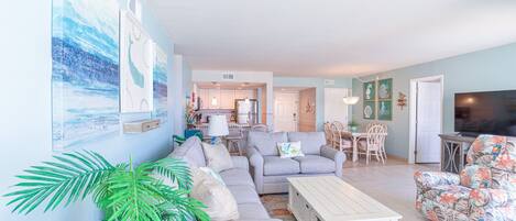 A spacious living room with a sectional sofa, a coffee table, and a TV. There is a dining area with a table and chairs, and a kitchen in the background.