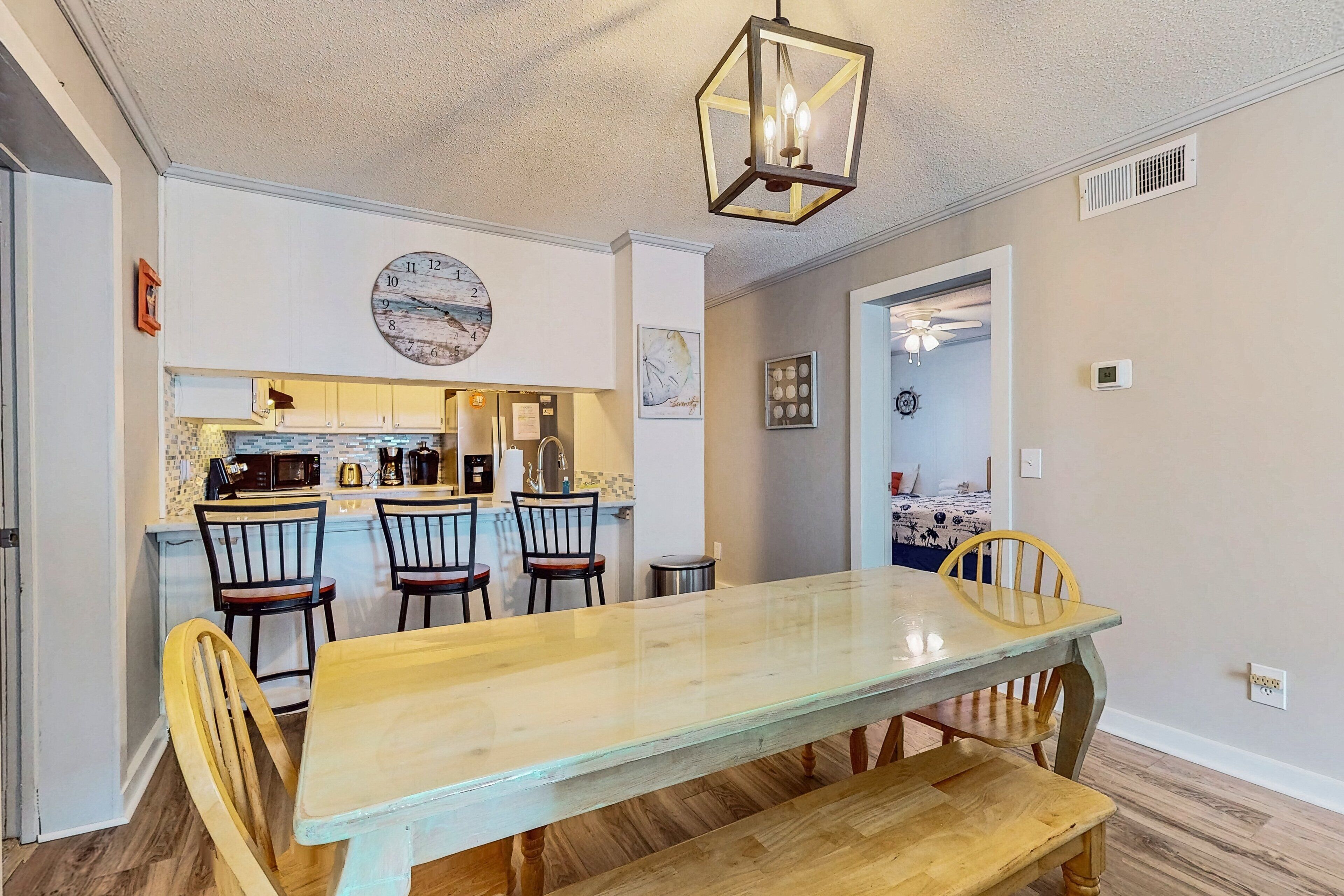 A dining area with a long wooden table and chairs, a kitchen in the background, and a clock on the wall.