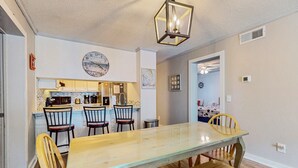 A dining area with a long wooden table and chairs, a kitchen in the background, and a clock on the wall. - Sandvillas C1: 2nd-Floor | Balcony | Pool (Ocean Isle Beach)