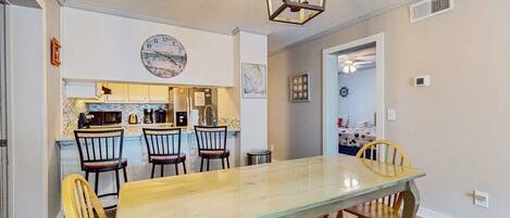 A dining area with a long wooden table and chairs, a kitchen in the background, and a clock on the wall.
