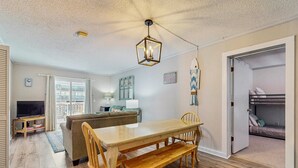 A cozy living area with a wooden dining table, benches, a sofa, and a TV on a stand. - Sandvillas C1: 2nd-Floor | Balcony | Pool (Ocean Isle Beach)