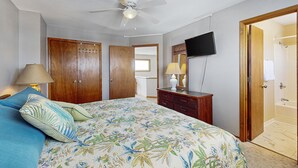 Une chambre à coucher avec un lit, une armoire en bois, un ventilateur de plafond, une télévision et une porte menant à la salle de bain.