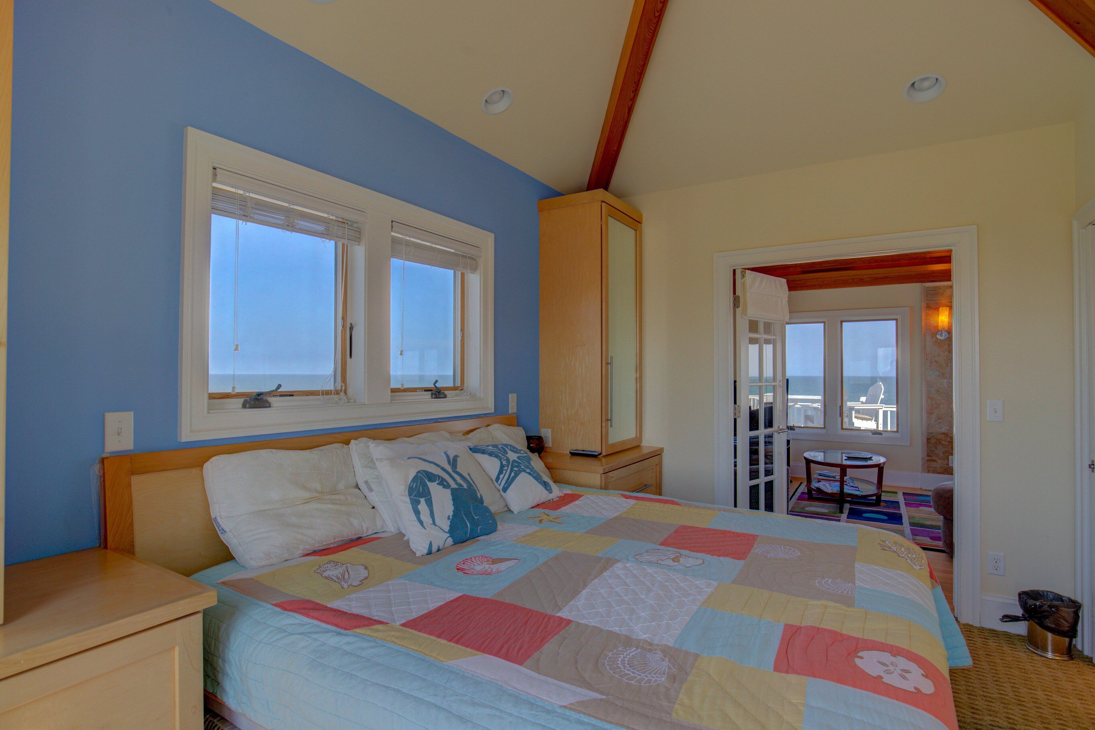 A bedroom with a large bed, two windows, a wooden ceiling, and a view of the outside.