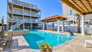 A swimming pool with a wooden deck and lounge chairs, surrounded by a multi-story building with balconies and a covered patio area. - Luxe ocean-view beach estate - amazing views, private pool, hot tub & elevator (North Topsail Beach)