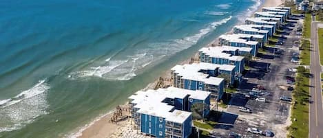 A coastal hotel with multiple blue buildings, a parking lot, and a beach.