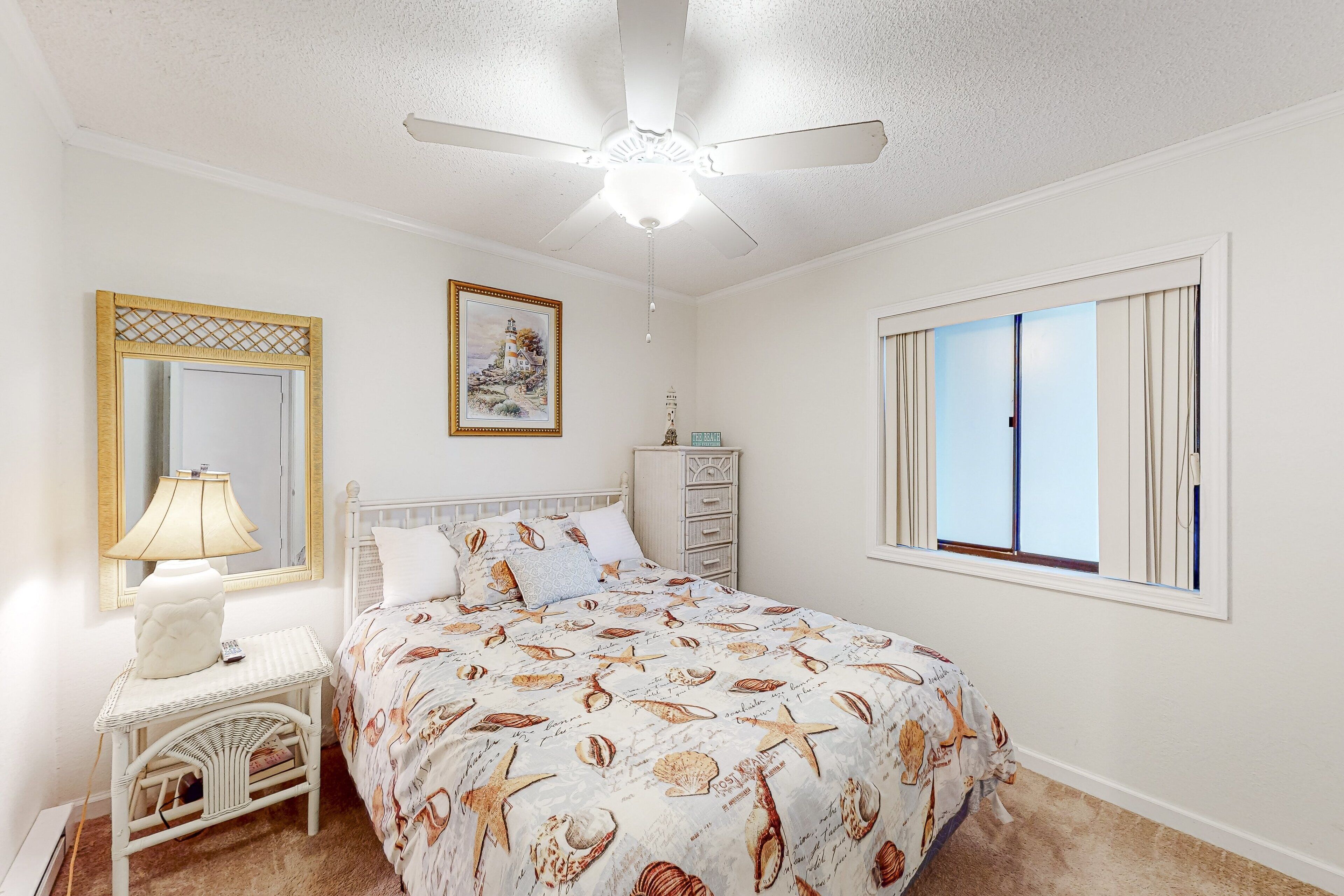 A bedroom with a bed, a nightstand, a lamp, a ceiling fan, a dresser, a window with curtains, and a framed picture on the wall.
