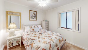 Una camera da letto con un letto, un comodino, una lampada, una ventola a soffitto, una cassettiera, una finestra con tende e un quadro appeso al muro.
