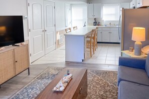 Una cocina moderna con armarios blancos, un comedor con sillas de madera, una isla central y un televisor de pantalla plana sobre un mueble de madera.