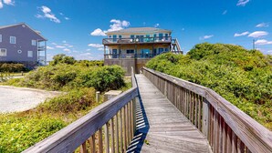 A wooden boardwalk leading to a multi-story building with balconies, surrounded by greenery. - Beachfront home with pool, private hot tub, beach access & lovely views (North Topsail Beach)