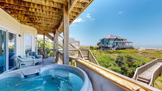 A hot tub area with a wooden deck, a covered seating area, and a view of a large house on a beach. - Beachfront home with pool, private hot tub, beach access & lovely views (North Topsail Beach)