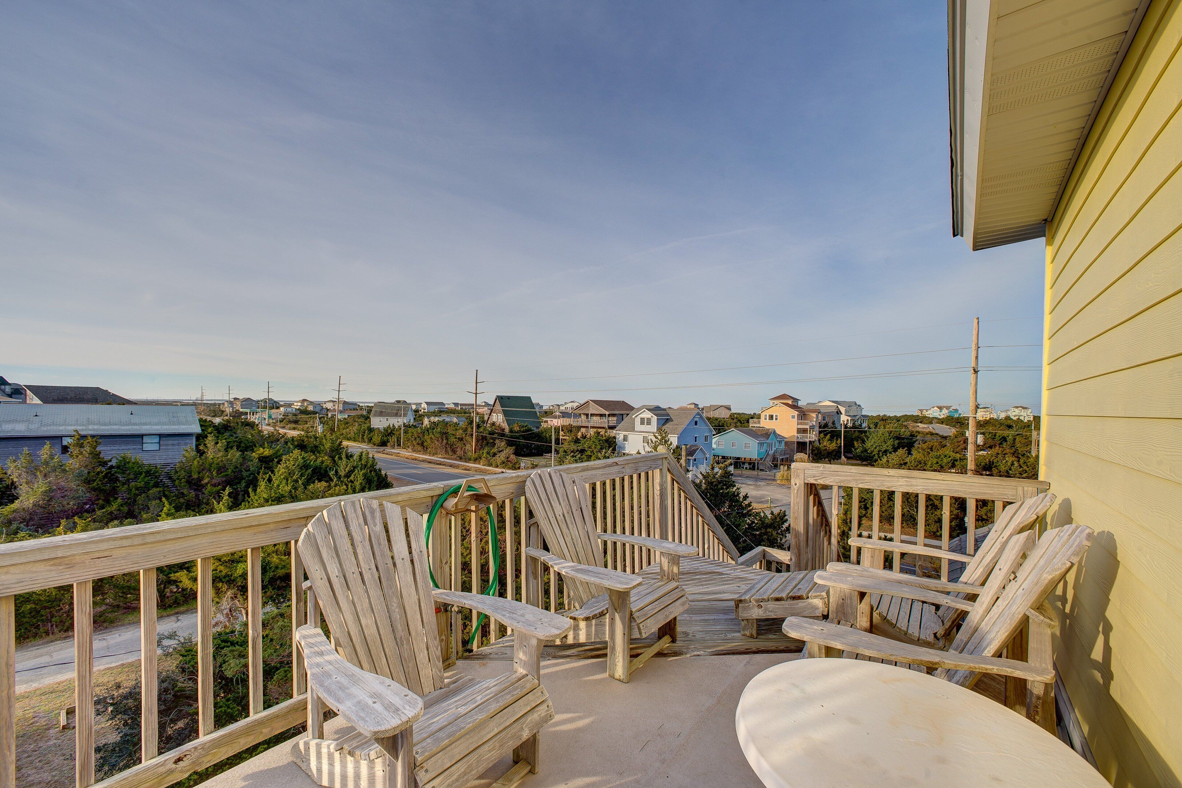 Um deck de madeira com cadeiras Adirondack e uma mesa, com vista para uma área costeira com casas e uma estrada.
