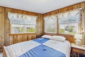 A bedroom with a wooden paneled wall, a bed with a blue and white quilt, two windows with white blinds, and a nightstand with a lamp. - Sandy Paws 394: Stunning Views, Pool Access, Pet-Friendly (Avon)