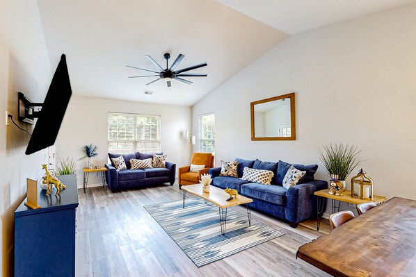 A spacious living room with a large sofa, a coffee table, and a dining table. There are two windows with blinds, a ceiling fan, and a mounted TV. - Family Tides- Sandpiper Bay: Shared pool (Sunset Beach)